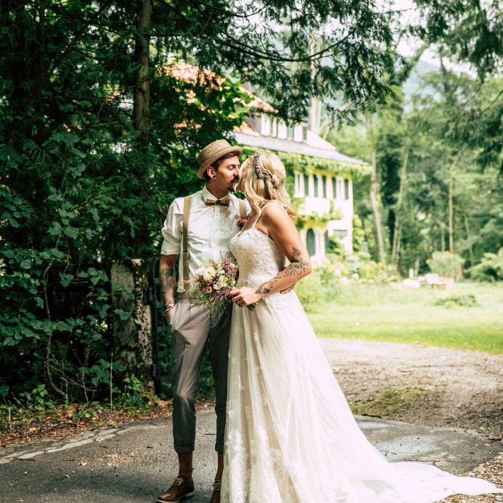 Ein Brautpaar küsst sich draußen auf dem Weg zu einem Haus, Schleppe, Brautstrauß, Hochzeit, Event Fotografie, WINGMEN Media