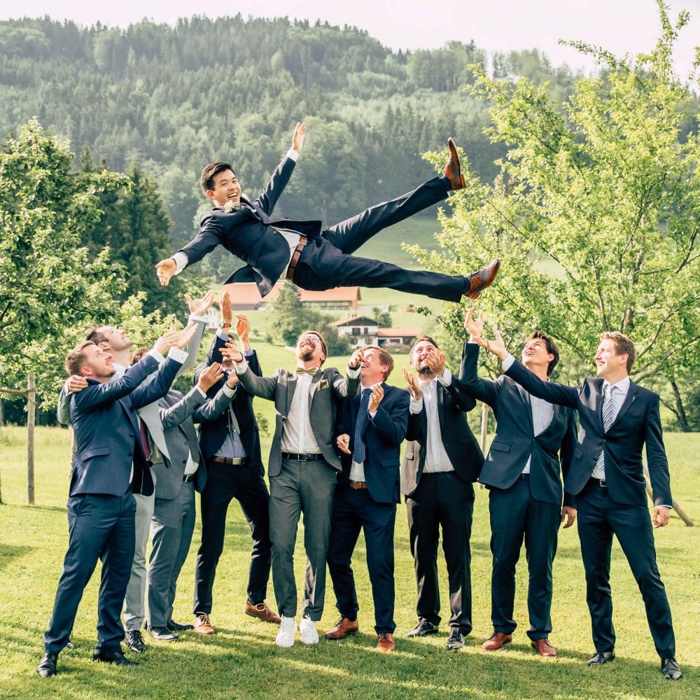 Ein Bräutigam wird von seinen Freunden hoch geworfen draußen auf einer Wiese, alle lachen, blauer Himmel, Event Fotografie, WINGMEN Media